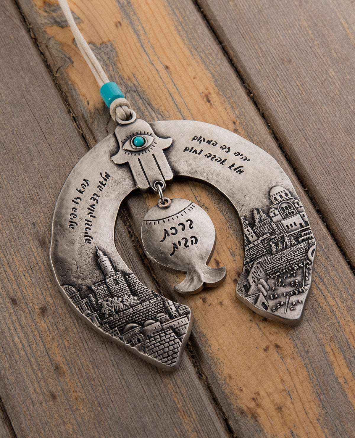 A unique and originally designed hanging Home Blessing ornament in the shape of a horseshoe. The horseshoe is embedded with an embossed image of Jerusalem on both sides. A pomegranate hangs from the center, with a Hamsa on top of it inlaid with a turquoise colored Swarovski crystal. The ornament is coated in sterling silver and decorated with words of blessing, love and light. Comes with a natural colored faux leather string decorated with a turquoise colored bead. Makes a great housewarming gift or an orig
