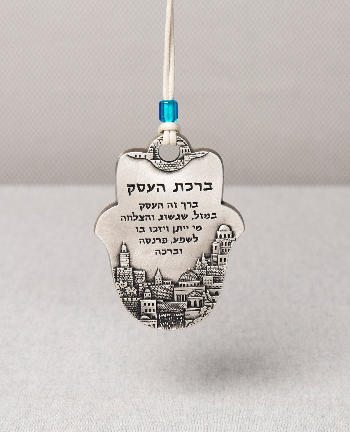 An impressive and excitingly designed hanging Hamsa Business Blessing. The Hamsa is coated in sterling silver and comes with a faux leather string for hanging decorated by a blue bead. Engraved on the bottom part of the Hamsa an image of beautiful Jerusalem, and in the upper part appear blessing words of abundance, luck, prosperity, success and good fortune. The hanging ornament makes the perfect gift as a gesture of good intention for any business, old or new. Length: 9 cm Width: 6 cm