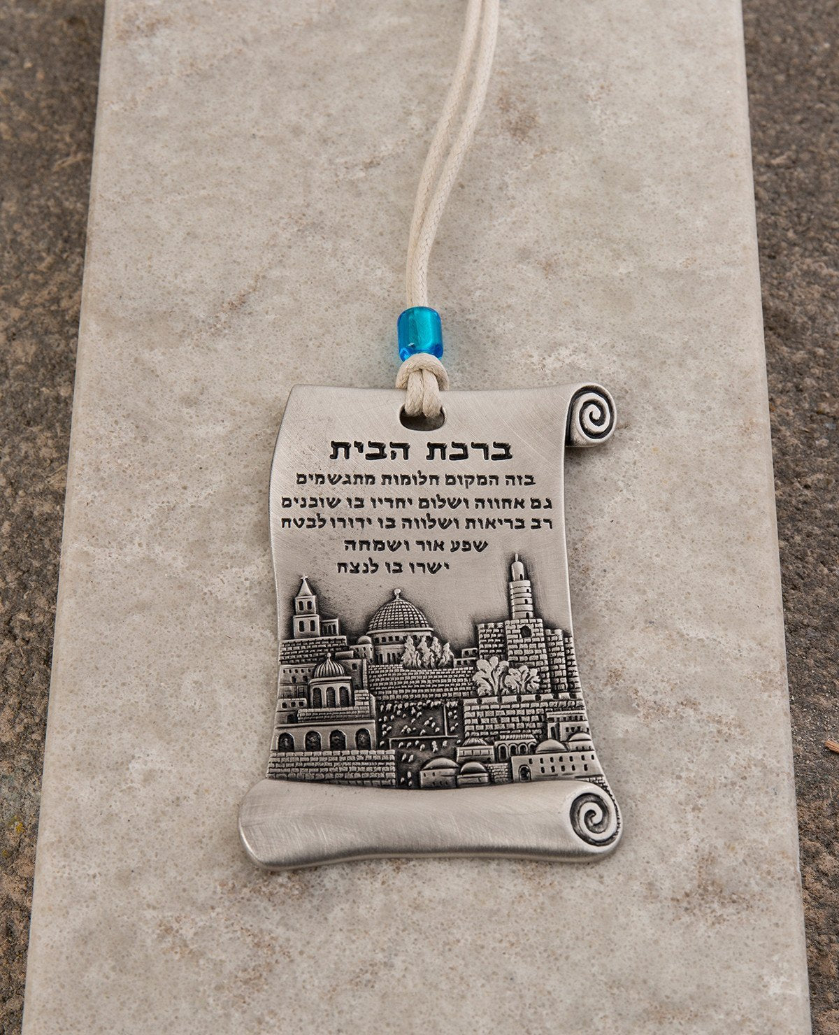 A hanging Home Blessing ornament in the shape of a scroll, coated in sterling silver and decorated with a beautiful embossed image of the city of Jerusalem. The scroll shaped design connects us back to ancient times, which already then the power of words were acknowledged as a force that can manifest abundance and joy in our life. The hanging ornament comes with a faux leather string decorated with a blue colored bead. Length: 9 cm Width: 5 cm