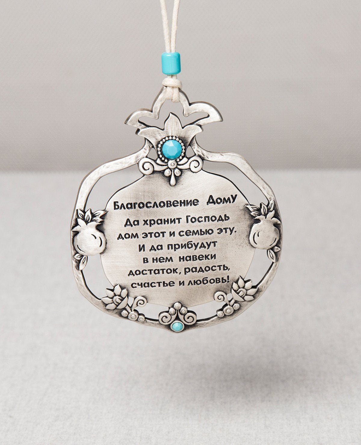 A hanging Home Blessing ornament in Russian. Designed in the shape of a pomegranate inside a pomegranate, coated in sterling silver and embedded with tuquoise colored Swarovski crystals. The outer layer of the pomegranate is adorned with flowers and little pomegranates. Comes with a faux leather string decorated with a turquoise colored bead. The pomegranate makes up one of the seven species, symbolizing abundance, fertility, beauty, and wisdom in different cultures. This charming hanging ornament will make