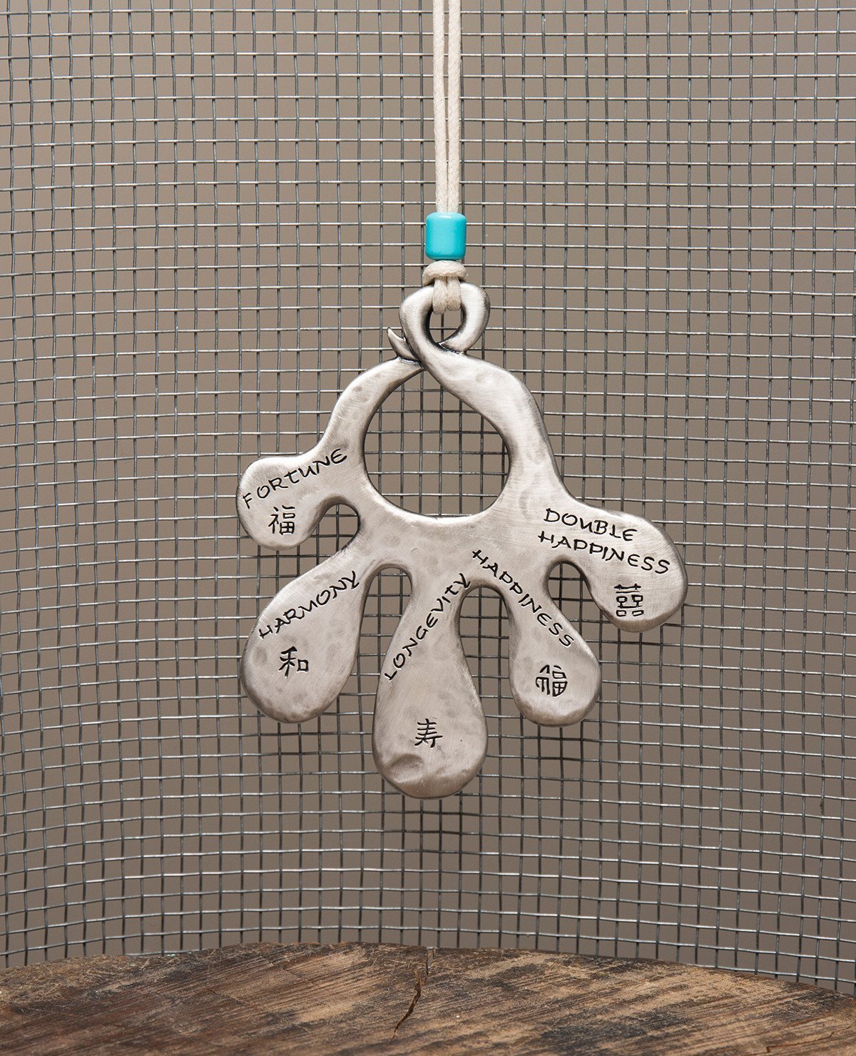 An open hand Hamsa wall ornament that hangs from a natural colored faux leather string decorated by a turquoise colored bead. The Hamsa is coated in sterling silver and each finger has a blessing engraved on it in English and Chinese. The blessings are from the world of Feng Shui, the ancient Chinese philosophy which represents the way of peace and harmony. Luck, abundance, harmony, peace, longevity, joy and more joy, these are the loving blessings which you can grant to whoever will receive this charming g