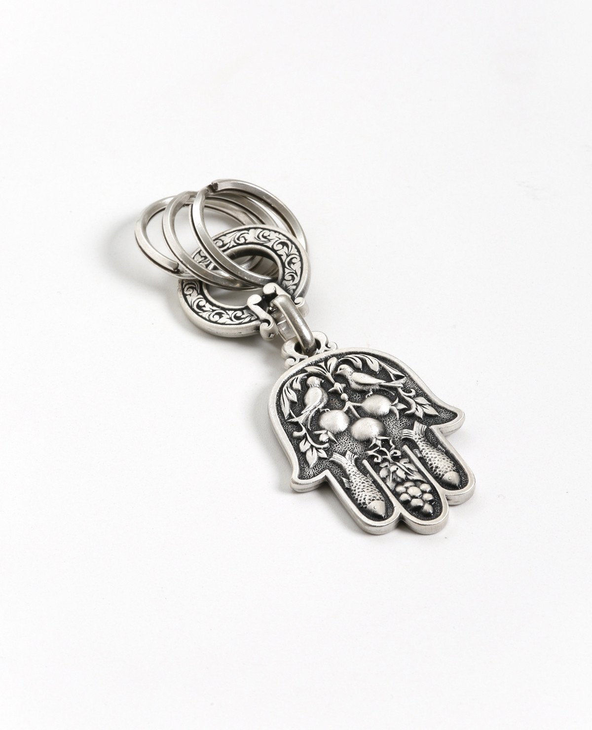 A double sided Hamsa keychain coated in sterling silver. On one side is a big eye embedded with a blue colored Swarovski crystal and embossed lions. On the other side are embossed birds, pomegranates, fish and grapes. The whole Hamsa is decorated in a filigree like style. The connecting links and the key rings are massive and strong. It's fun to connect our most important keys to it and most definitely to grant our loved ones with such a beautiful Hamsa, blessed with the many icons of good fortune, protecti