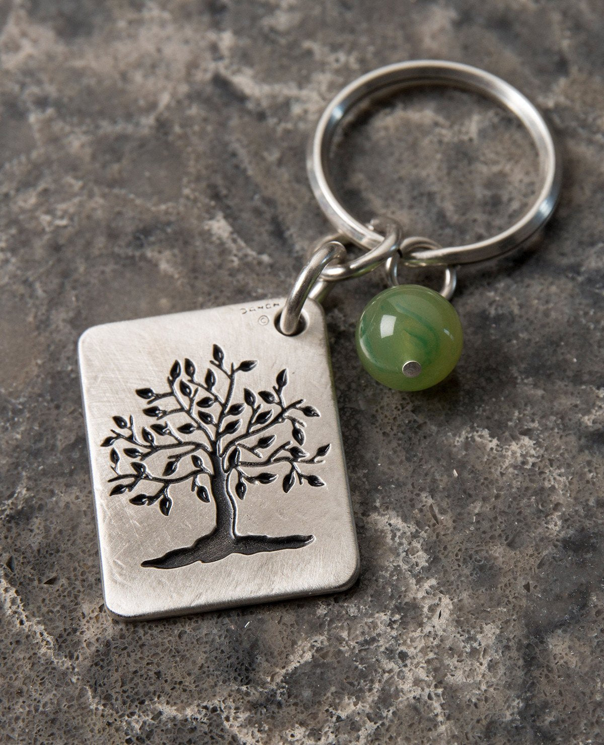 A keychain that is all charm, beauty and carries a very human message. Designed as a rectangular plate with the words "Be Kind Be You Be True" written in English on one side. The other side has an embossed image of a wide treetop and roots. Next to the plate hangs a stunningly beautiful green stone. The keychain is coated in sterling silver and is strong and reliable. A great gift for him or for her, here or anywhere, with the deepest and simplest message: be generous, be who you are, be real. This signific