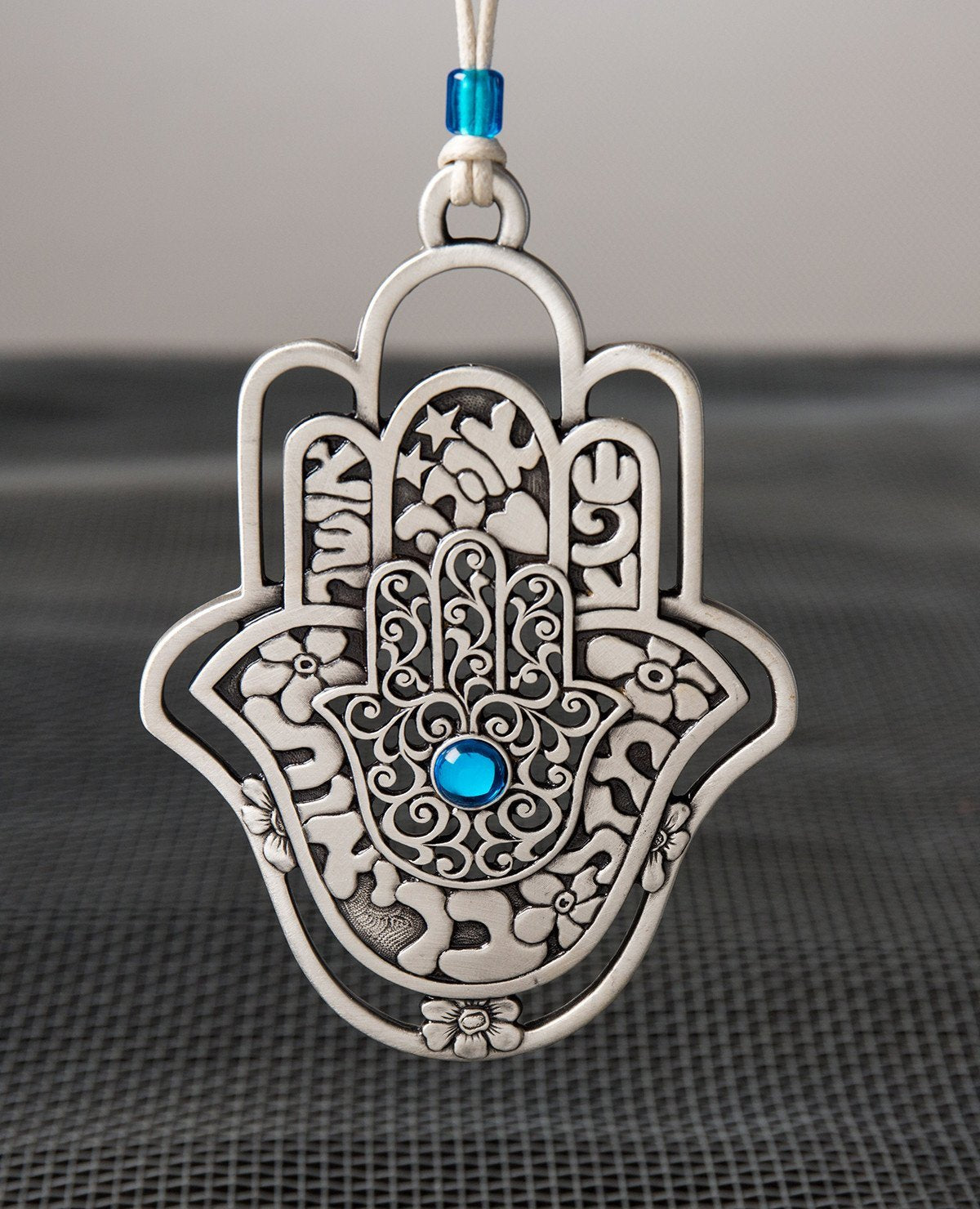 A hanging Hamsa ornament, designed in exquisite beauty: a Hamsa within a Hamsa within a Hamsa. The outer one - a hollow frame decorated by delicate flowers. The middle one - all embossed words of blessing - for abundance, luck, health, happiness and love. The inner one - filigree decoration with a blue stone at the center. The ornament is coated in sterling silver and comes with a faux leather string deocorated by a blue bead. An impressive and very meaningful gift that is a pleasure to grant to our loved o
