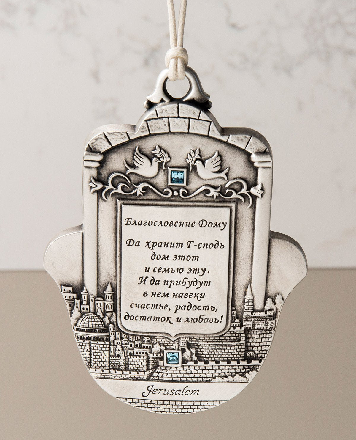 An impressively designed and uniquely decorated Hamsa hanging ornament coated in sterling silver. Decorated with an embossed image of Jerusalem and a pair of peace doves with an olive branch, alongside blessing words of love, joy, laughter and light for the home. Makes a thoughtful and loving gift that will brighten up any house. The Hamsa is embedded with azure Swarovski crystals and a natural colored faux leather string. 
Additional languages available: English and Hebrew.  Length: 13 cm  Width: 9 cm