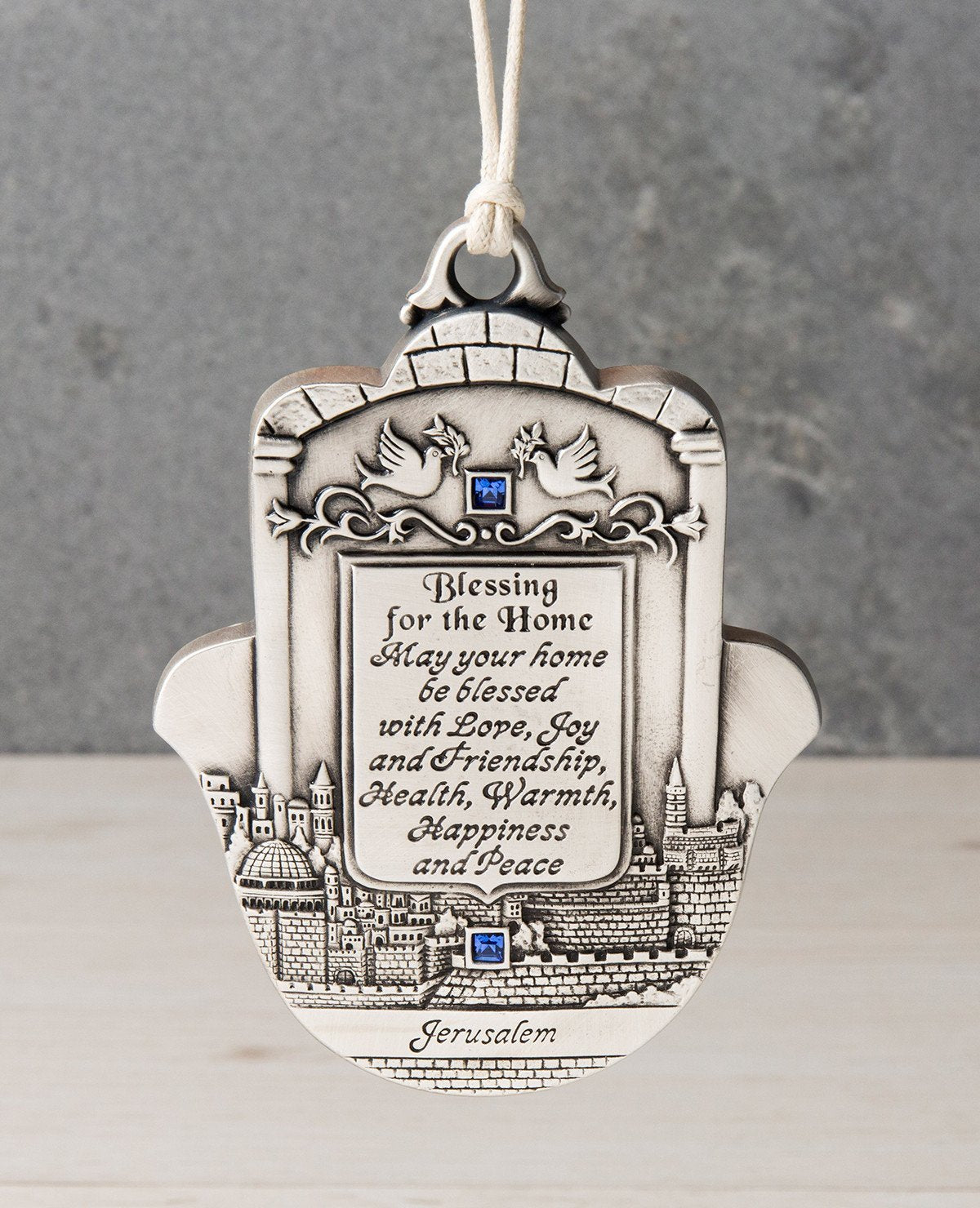 An impressively designed and uniquely decorated Hamsa hanging ornament coated in sterling silver. Decorated with an embossed image of Jerusalem and a pair of peace doves with an olive branch, alongside blessing words of love, joy, laughter and light for the home. Makes a thoughtful and loving gift that will brighten up any house. The Hamsa is embedded with blue Swarovski crystals and a natural colored faux leather string. 
Additional languages available: Hebrew and Russian.  Length: 13 cm  Width: 10 cm