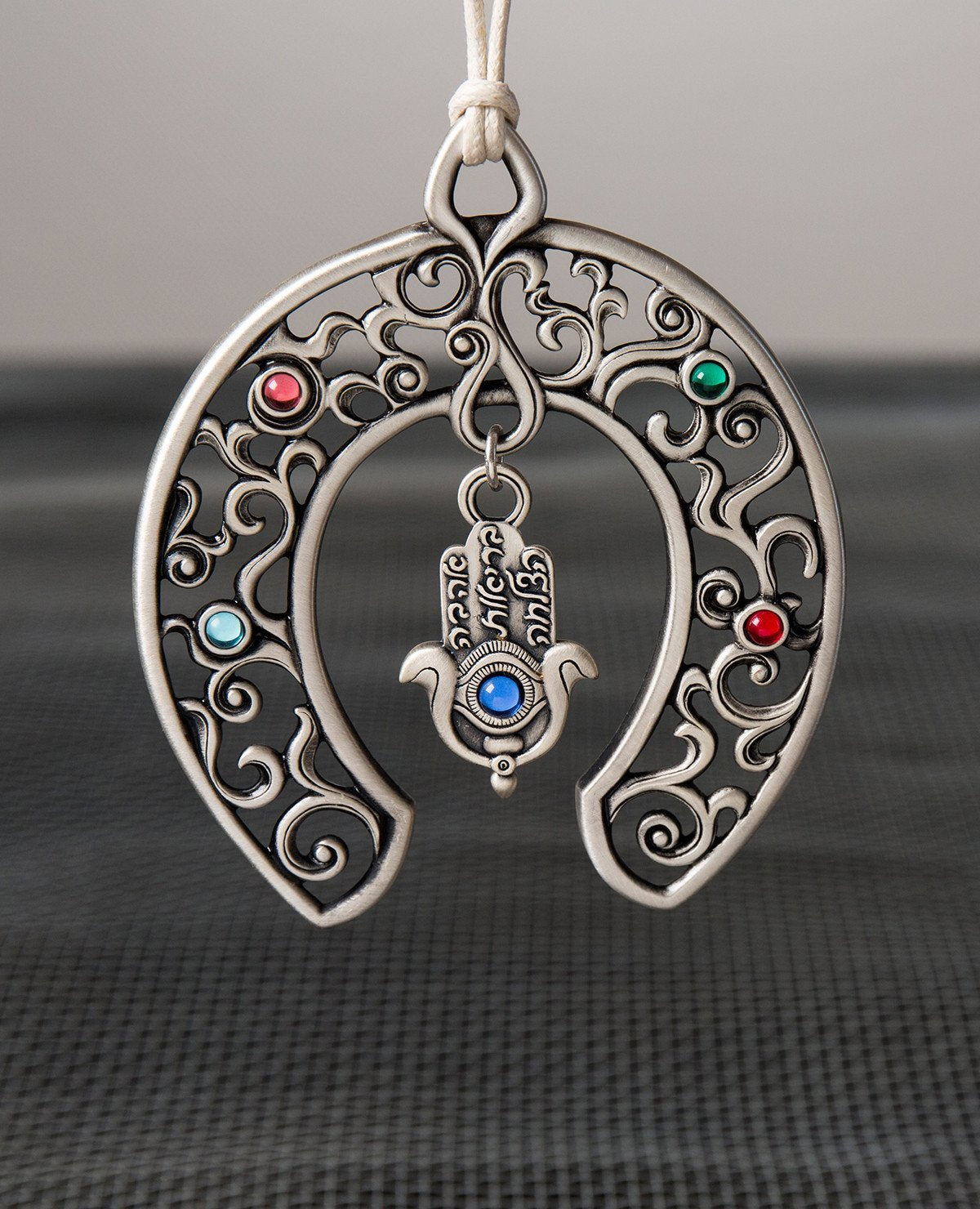 An astonishingly designed hanging horseshoe wall ornament: a large horseshoe, decorated by filigree and embedded with colorful stones. At its center hangs a designed Hamsa, which has an engraved eye on it embedded with a blue stone and the words "love", "health" and "luck". The ornament is coated in sterling silver and comes with a natural colored faux leather string for hanging. The horseshoe and the Hamsa are both well known charms to ward off the "evil eye", and also bring blessings of abundance and good
