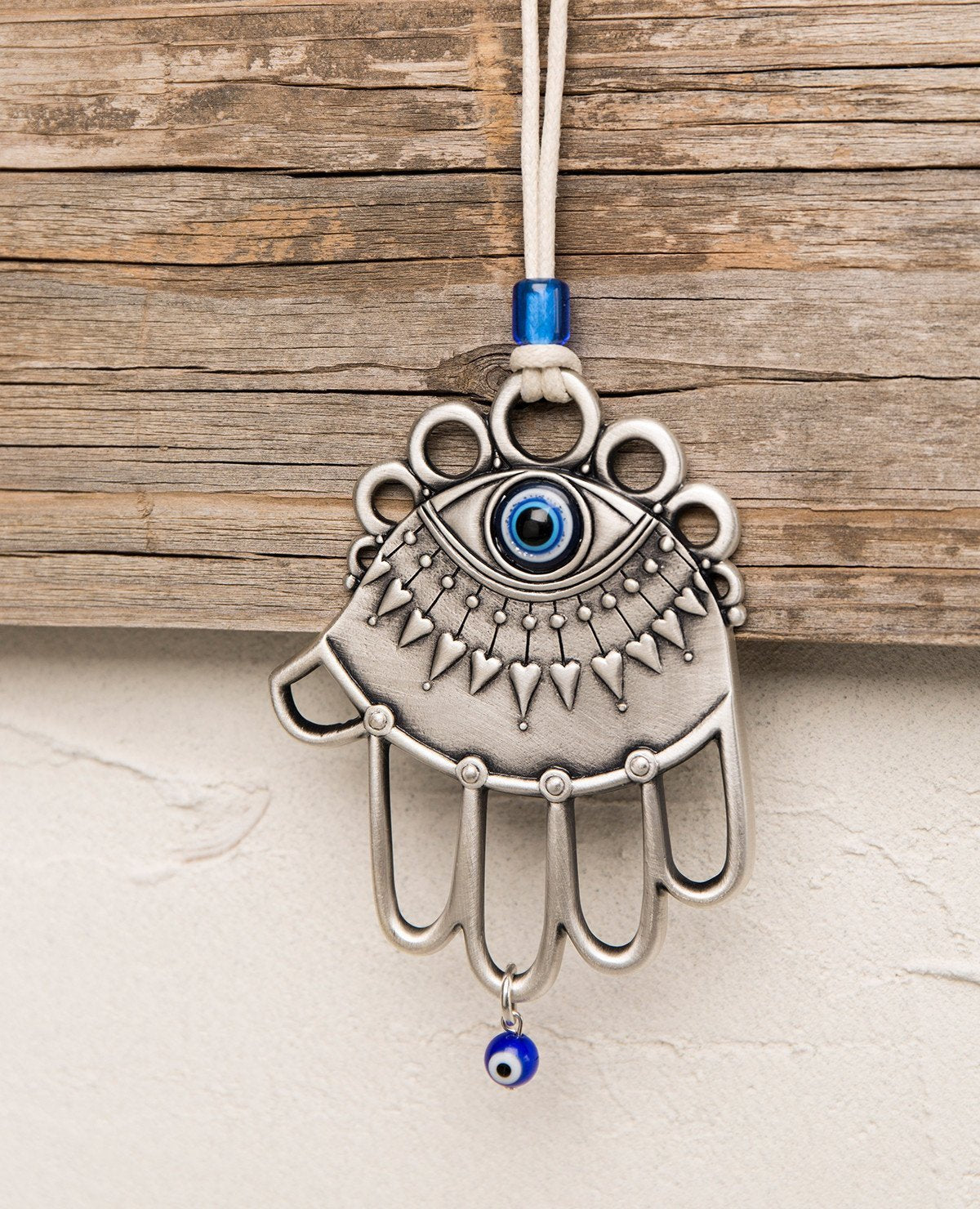 A one of a kind hanging Hamsa ornament. It is designed with a combination of different styles, some of which are hollow, while others are solid decorated surfaces. Embedded at the top of the Hamsa is a large blue eye, decorated by a lower eyelash in the shape of heart arrows. The fingers of the Hamsa are hollow and at their bottom hangs a small blue eye. The ornament is coated in sterling silver and comes with a faux leather string for hanging, decorated by a blue bead. 
The blue eye is considered a charm a