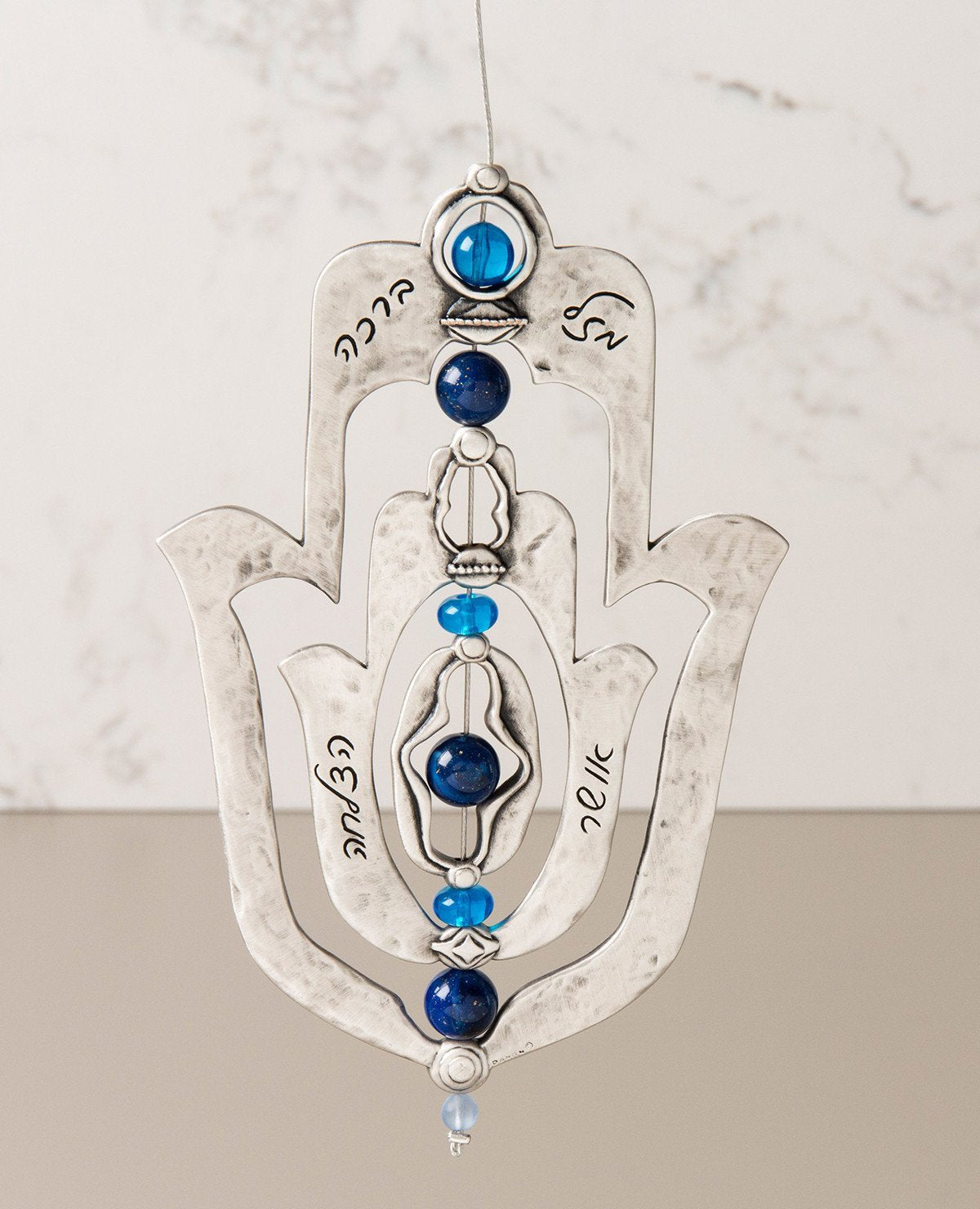 An impressive and neatly designed hanging Hamsa ornament coated in sterling silver. Set on an axis inside a large Hamsa is a smaller Hamsa. The axis is made from a thin string coated in silver and decorated by blue beads. The ornament spins on the axis, and on each side appear different words of blessing. The qualities of the Hamsa on one side and the power of the words respectively on the other. A great gift that is also a joyful and heartwarming blessing, for any occasion. The ornament comes with a faux l