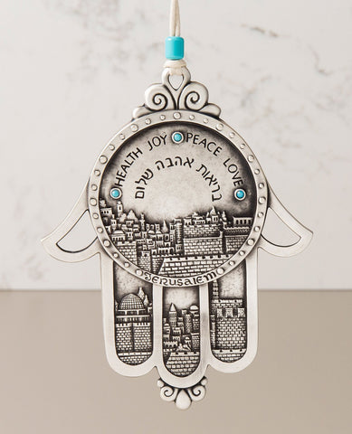 Wow! What an extraordinarily beautiful gift! A hanging Hamsa ornament coated in sterling silver. The body of the Hamsa along with the three middle fingers are all decorated by an embossed image of the stunning views of Jerusalem. Engraved on the Hamsa in Hebrew and English are the words: "Health, Love and Peace" and in English the word "Jerusalem". The Hamsa is embedded with turquoise colored Swarovski stones and comes with a natural colored faux leather string for hanging. The string is decorated with a tu