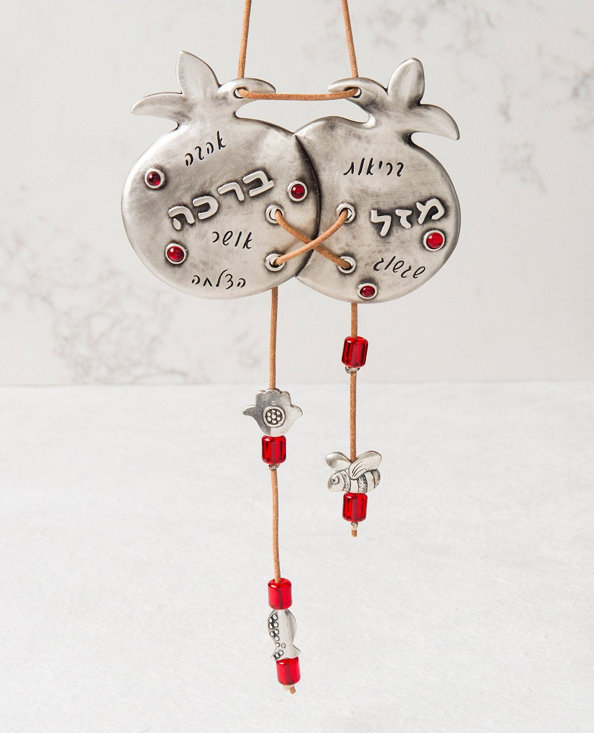 Two are better than one! A sweet and charming hanging ornament with a smile and a blessing. The pair of pomegranates are connected to each other, coated in sterling silver and embedded with red crystals. A natural colored faux leather string is weaved between the pomegranates, with the ends dangling from the bottom of each, decorated by red beads and tiny heart and Hamsa decorations. The top part of the string serves for hanging the ornament. Engraved on the pomegranates are words of blessing: luck, health,