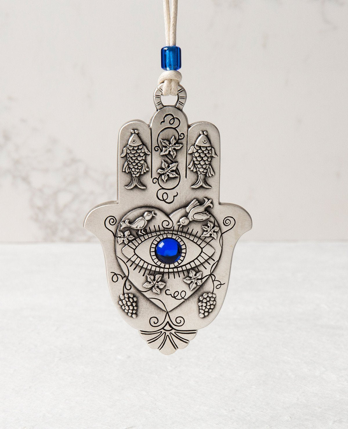 A hanging Hamsa ornament packed with goodness! The Hamsa is coated in sterling sivler and comes with a natural colored faux leather string for hanging, decorated by a blue bead. At the center of the Hamsa is a big heart with an embossed eye inside embedded with a blue colored stone (against the "evil eye"). Above the eye are chirping birds (of peace and love). The Hamsa is decorated with embossed fish (for luck), as well as vine leaves and grape clusters (for abundance). What else could we ask for? Just the