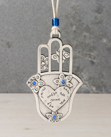 An aritistic, charming and delicately designed Hamsa hanging ornament. The Hamsa is coated in sterling silver and on its bottom part has a smooth plate with a big heart on it. Written inside the heart are seven blessings for: love, abundance, success, happiness, health, joy and luck. Surrounding the heart are flower decorations embedded with blue colored stones. On the top part of the Hamsa are three hollowed out fingers, with butterflies in between them. The ornament comes with a natural colored faux leath