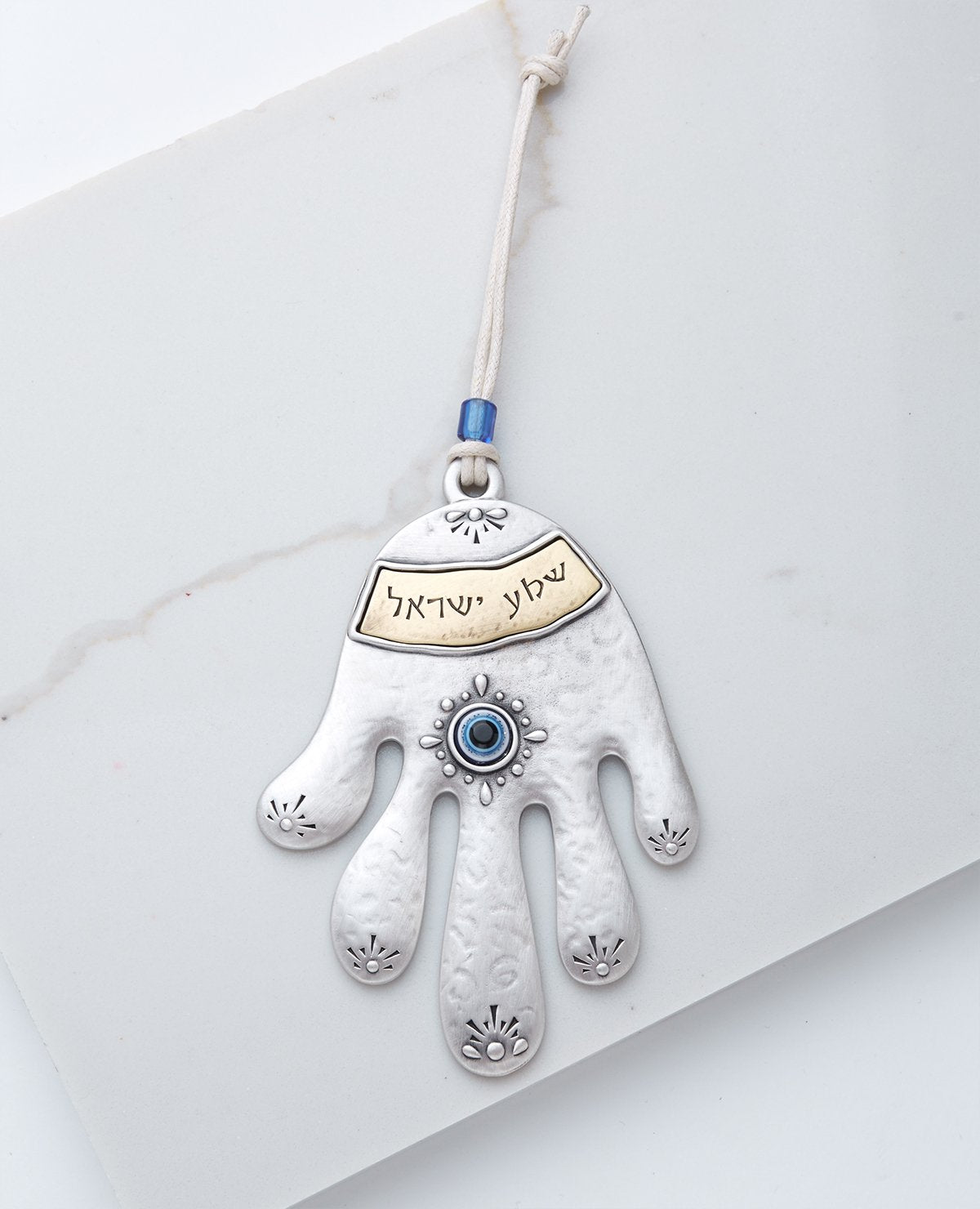 A charming and inspiring Hamsa hanging wall ornament, coated in sterling silver and brass. It hangs from a natural colored cotton string decorated with a blue bead. The rich and stylish design and the word of "Shma Israel" in Hebrew make a wonderful gift that contains beauty and protection. Grant it with love (and don't forget to spoil yourselves as well). Length:14 cm Width: 10 cm