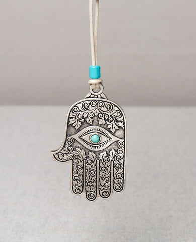 A hanging Hamsa ornament decorated with a filigree engraving, coated in sterling silver and embedded with a turquoise colored stone. The Hamsa hangs from a leather string which is decorated by a turquoise colored bead. The Hamsa is a well known good luck charm that contains qualities that protect from the "evil eye". That's why it makes such a sweet and joyful gift to hang on the wall, the car mirror, and in any place where we ask to grant protection and a good blessing.  Length: 9 cm  Width: 5 cm