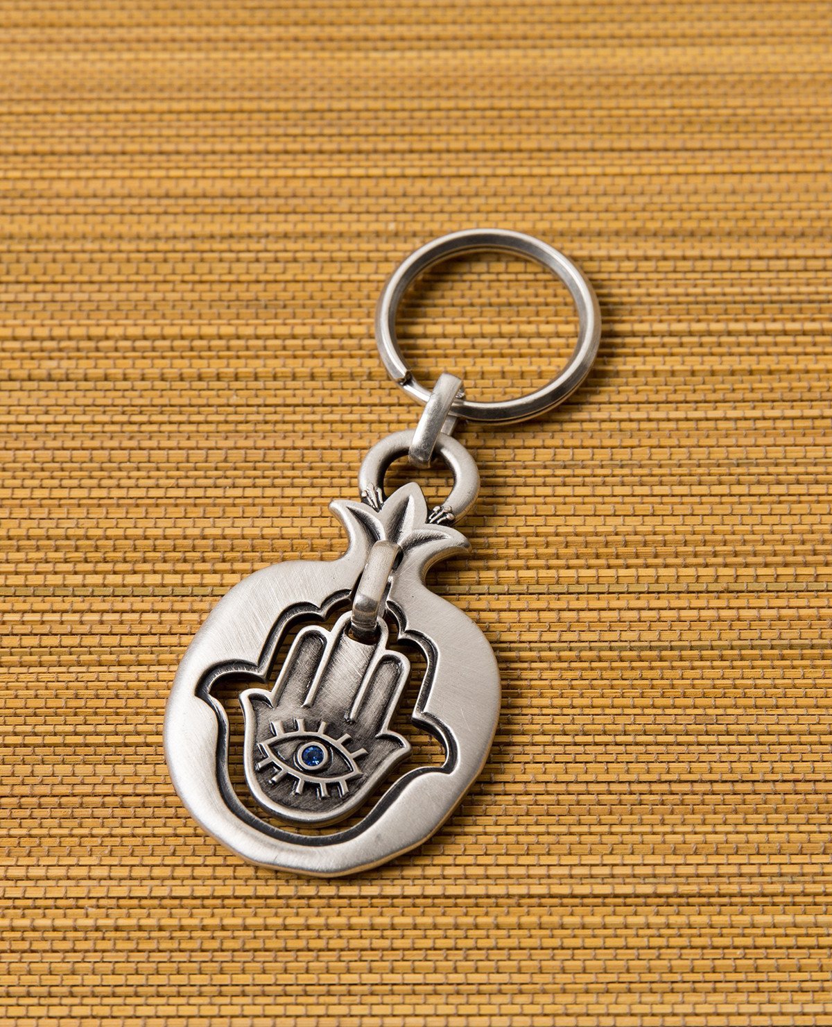 A blessing every time you open and lock. A charming keychain coated in sterling silver and carrying a blessing that follows you wherever you go. A Hamsa hangs within the hollow pomegranate frame. On it's one side an eye embedded with a blue colored crystal and a charm against the "evil eye", and on the other side appear the words "Hamsa On You" decorated with flowers, for luck and blessing. All of these are contained within the pomegranate, which brings the blessings of abundance and fertility. A suitable a
