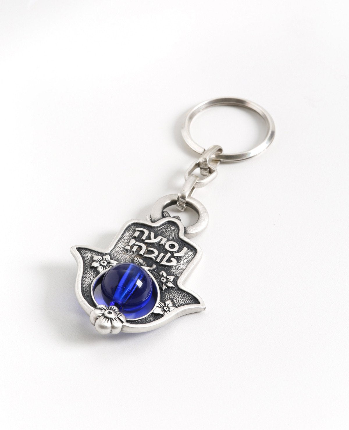 A delicate and charmingly designed Hamsa keychain. The Hamsa is coated in sterling silver and has a stunning blue bead which can be seen from both sides of the Hamsa threaded on its bottom part. On the one side is a delicate flower decor with the embossed words "Bon Voyage!" while the other side is decorated with flowers and a pair of fish. The keychain is strong and reliable. Makes a great present for anyone whose vehicle's keys are in need of a pretty holder. Blessed with the Hamsa charm against the "evil