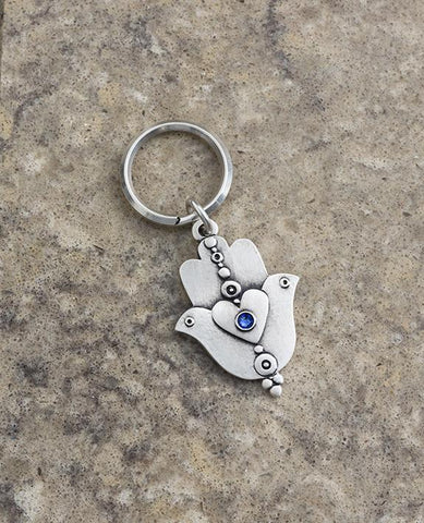A extraordinarily beautiful keychain with a rare design, coated in sterling silver. On one side of the Hamsa is a dove inlaid with a blue colored Swarovski crystal. Engraved on top is the Kohen's blessing "May the Lord bless you and keep you safe". On the other side of the Hamsa are two doves looking in opposite directions, with a heart in between them that is also embedded with a blue stone. A keychain that is all beauty and blessings. The Hamsa's blessing for good luck, the doves blessing for peace and lo