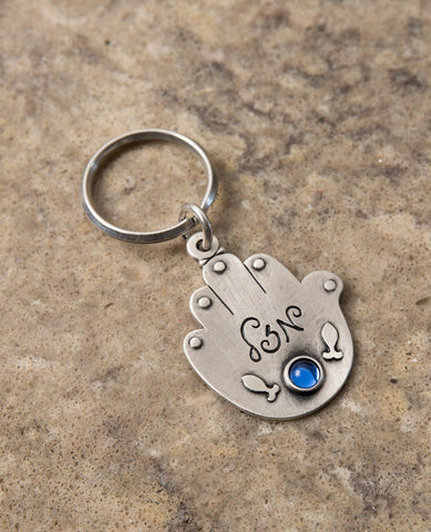 An exciting double-sided Hamsa keychain. On one side of the Hamsa the words "Good journey and safe travels!" are written. Written on the other side is the word "Luck" with a blue stone embedded underneath, and next to it an embossed image of two fish.
The Hamsa is coated in sterling silver and is strong and reliable to accompany your loved ones throughout the entire journey. A charming and loving gift for anyone about to embark on a trip in the voyage of life. A present that will leave our loved ones with a