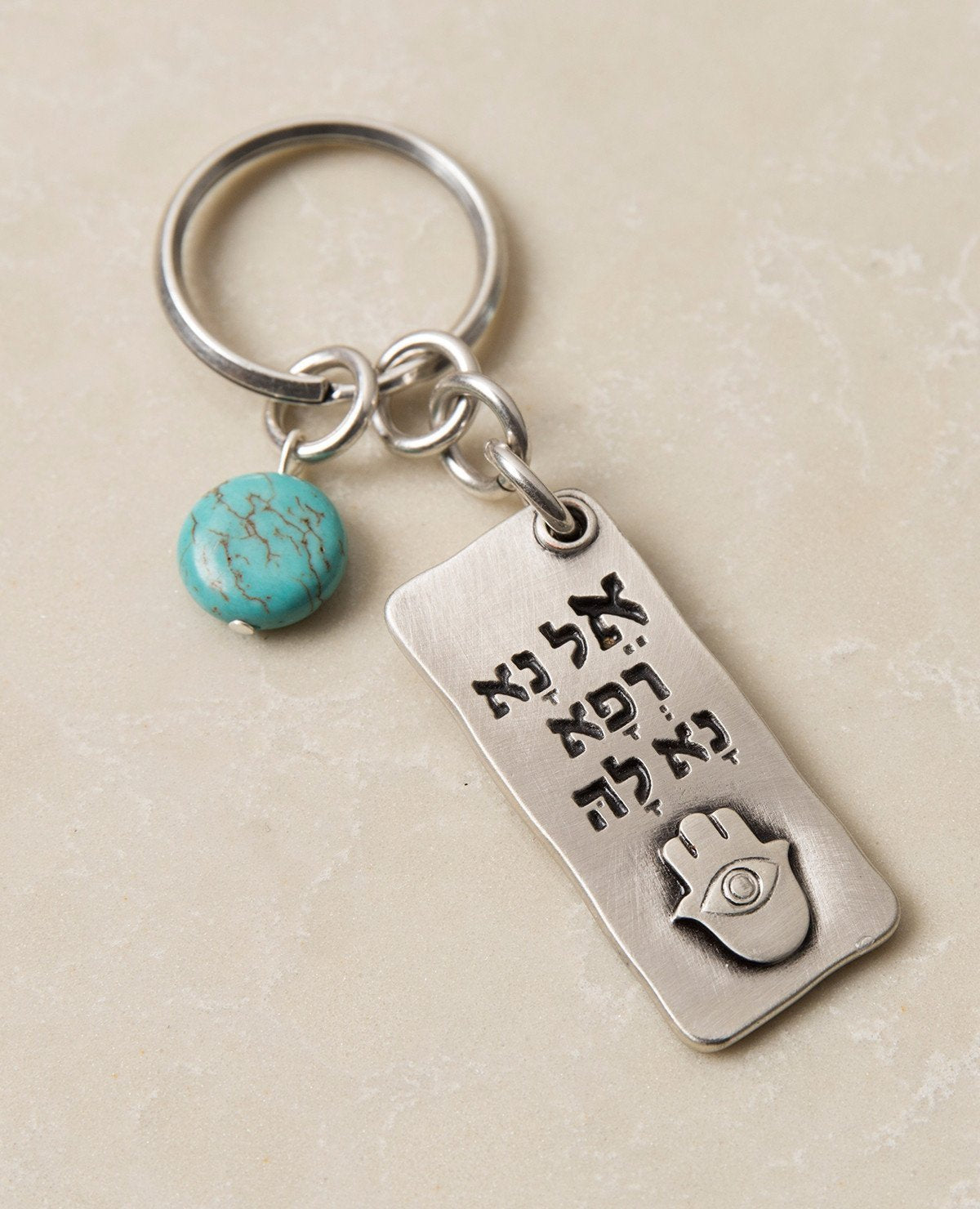 A beautiful and excitingly designed keychain, the content of which was taken from a prayer by Moses for the health of his sister, and it is in fact a prayer for good health. Shaped as a rectangular plate that has the sentence "Please, God, heal her" written in Hebrew on one side, and underneath an embossed image of a Hamsa with an eye. The other side contains blessings, in both English and Hebrew, for quick recovery, good health, and longevity. Next to the plate hangs a pretty turquoise stone. The keychain 