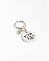 The freedom to be exactly who you are! An authentic and captivating keychain that you should always have ready to hand to someone special. The keychain is designed as a circle with the words "Freedom is a state of mind" written on one side, and on the other a drawing of a pair of birds perched on a tree branch. The circle is connected to the keyring with three strong rings. The top one of the three has charming green and orange beads hanging from it. Simple, precise, beautiful and oh so significant. The key