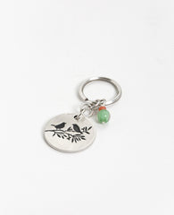 The freedom to be exactly who you are! An authentic and captivating keychain that you should always have ready to hand to someone special. The keychain is designed as a circle with the words "Freedom is a state of mind" written on one side, and on the other a drawing of a pair of birds perched on a tree branch. The circle is connected to the keyring with three strong rings. The top one of the three has charming green and orange beads hanging from it. Simple, precise, beautiful and oh so significant. The key
